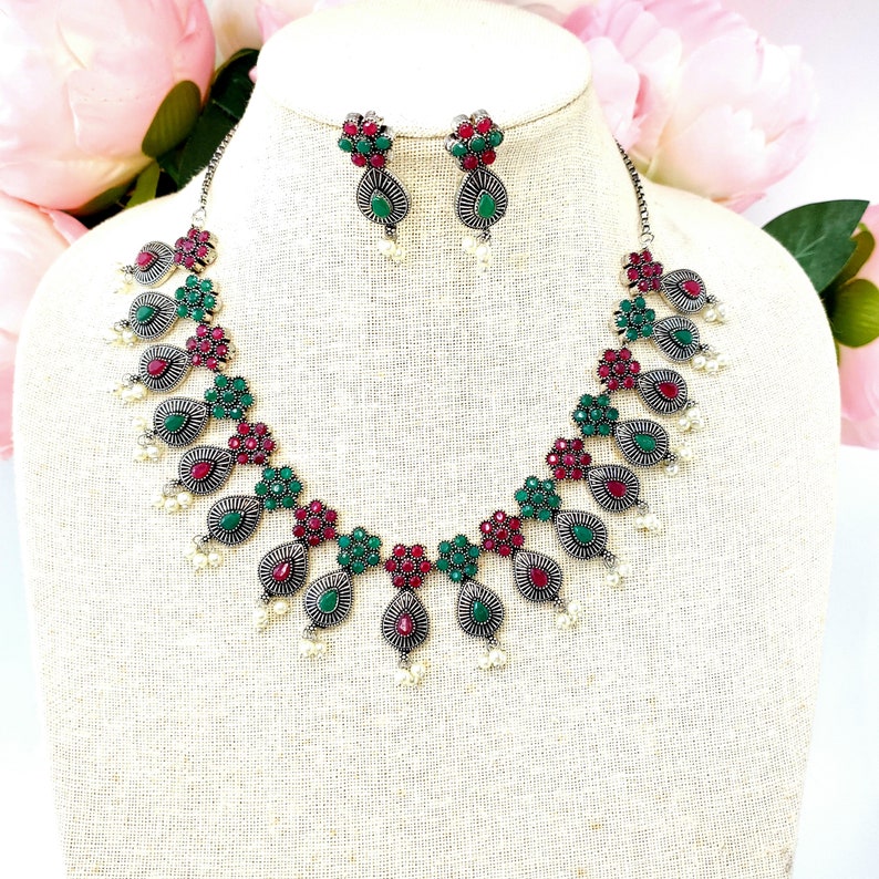 May include: A silver necklace and earrings set with red and green gemstone accents. The necklace features a teardrop design with pearl accents.