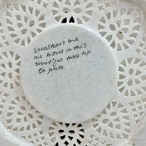 Puede incluir: Mesa de m&aacute;rmol blanco con intrincados dise&ntilde;os recortados. La parte posterior de la mesa tiene una nota escrita a mano que dice: "2000 piedras est&aacute;n incrustadas en esta hermosa mesa.  Do plate."