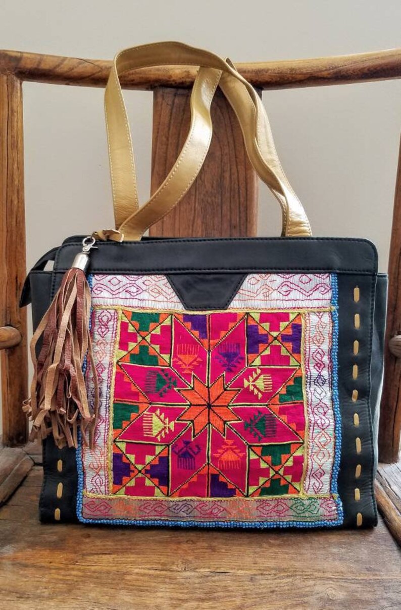 May include: Black tote bag with gold handles and a colourful embroidered front panel. The panel features a geometric starburst design in shades of pink, orange, green, and purple. A brown leather tassel hangs from the zipper.