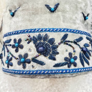 May include: A white fabric with a blue and silver embroidered floral design. The design features a large flower with a blue center and smaller flowers with turquoise centers. The fabric is trimmed with a blue and silver ribbon.
