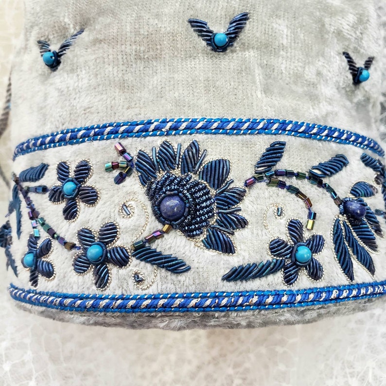 May include: A white fabric with a blue and silver embroidered floral design. The design features a large flower with a blue center and smaller flowers with turquoise centers. The fabric is trimmed with a blue and silver ribbon.