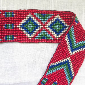 May include: Red beaded trim with a geometric pattern in blue, green, and white. The trim is made of small, square beads.