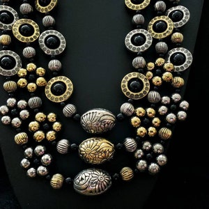 May include: A multi-strand necklace with black, gold, and silver beads. The necklace features round and oval beads with intricate designs.