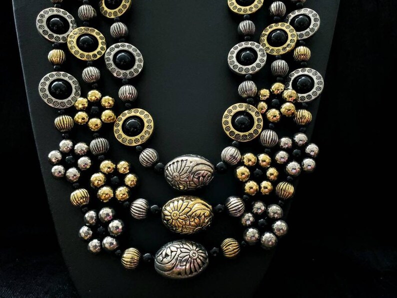 May include: A multi-strand necklace with black, gold, and silver beads. The necklace features round and oval beads with intricate designs.