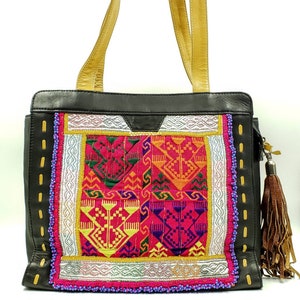 May include: Black leather tote bag with gold-coloured handles and a colourful embroidered front panel. The panel features geometric patterns in pink, yellow, green, and orange, with silver and gold accents. A brown tassel hangs from the side.