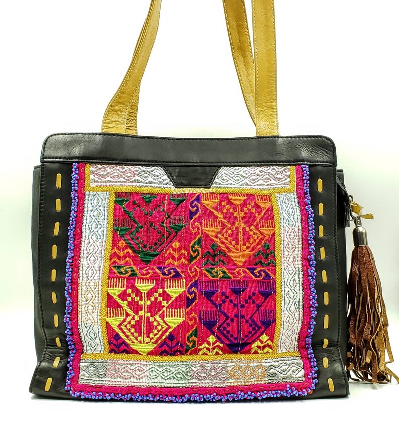 May include: Black leather tote bag with gold-coloured handles and a colourful embroidered front panel. The panel features geometric patterns in pink, yellow, green, and orange, with silver and gold accents. A brown tassel hangs from the side.