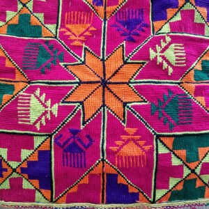 May include: Close-up of a colourful textile with a geometric pattern. The design features a central orange star surrounded by pink, green, purple, and yellow shapes. The edges are trimmed with a decorative border and blue beads.