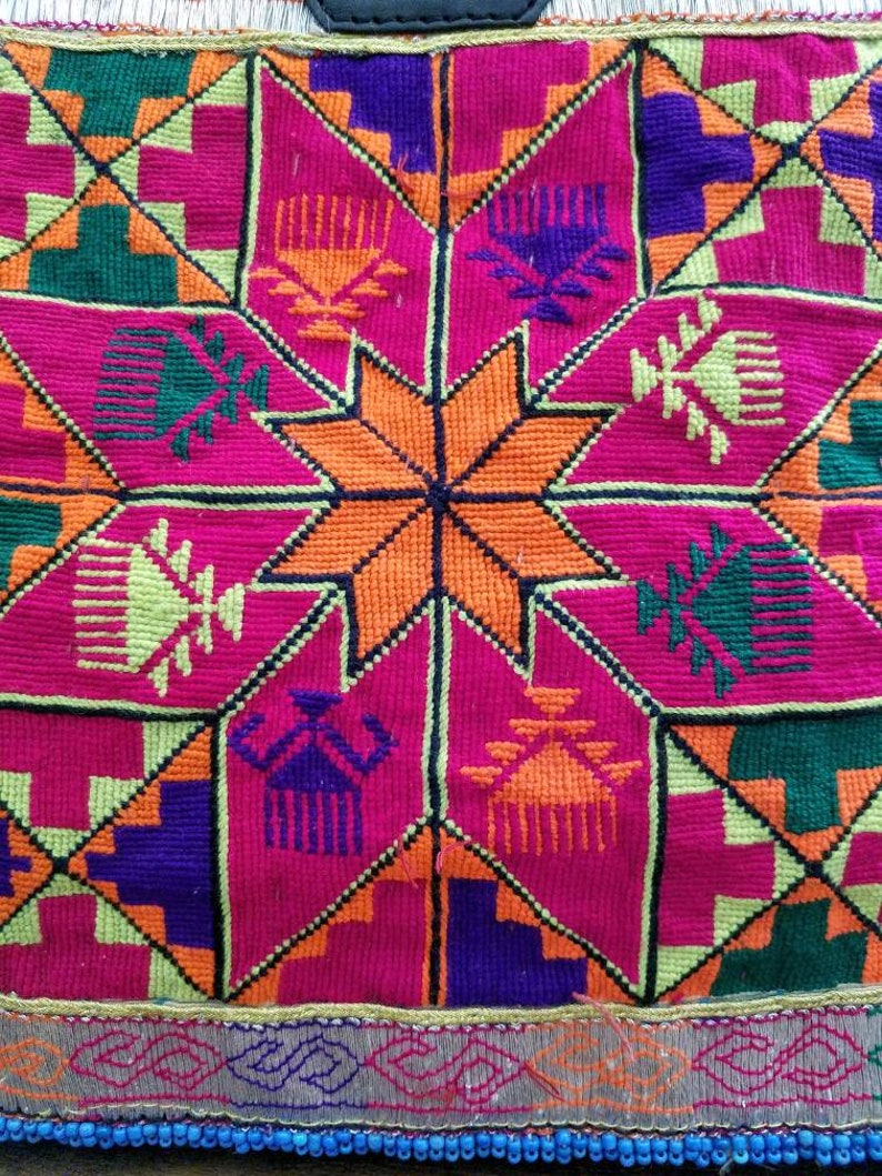May include: Close-up of a colourful textile with a geometric pattern. The design features a central orange star surrounded by pink, green, purple, and yellow shapes. The edges are trimmed with a decorative border and blue beads.