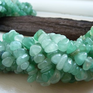 May include: A strand of light green gemstone beads, each bead is a smooth, irregular shape, resembling a small pebble. The beads are arranged in a loose, flowing pattern, creating a natural and organic look.