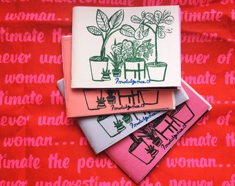 May include: A stack of small square zines with green plant illustrations on a red background. The top zine is white, with others in pink, peach, and lavender. The background is red with pink text that reads "Never underestimate the power of a woman..."