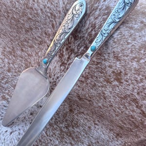 May include: A silver-toned cake server and a serrated knife set. Each handle features intricate engraved scrollwork patterns and a single small, round, bright blue turquoise stone inlay set near the base of the metal blade.