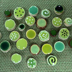May include: A collection of small, round ceramic buttons in various shades of green. The buttons feature diverse surface designs, including solid colors, white polka dots, white swirls, and radial patterns, all with a glossy, smooth finish.