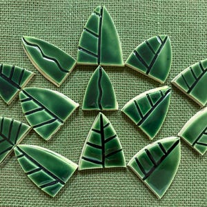 Puede incluir: Una colección de trece piezas de mosaico cerámico con forma de hoja en varios tonos de verde brillante. Cada pieza presenta una superficie esmaltada y lisa con líneas oscuras y hundidas que forman un patrón de nervaduras.
