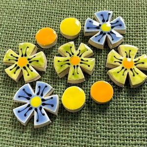May include: A collection of ceramic flower and circle tiles in various colors. The flower tiles have yellow, green, and blue petals with black accents and yellow centers. The circle tiles are yellow and orange. The tiles are arranged on a green textured surface.