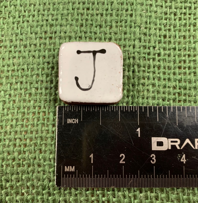 May include: White ceramic tile with a black handwritten letter J. The tile is about 1 inch square.
