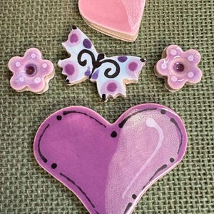 May include: Decorative ceramic pieces in heart, flower, and butterfly shapes. The pieces have a glossy finish in pink, purple, and blue hues, set against a textured green background. Measurements are not available.