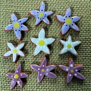 May include: A collection of nine ceramic star-shaped buttons arranged in three rows. The buttons feature glossy finishes in shades of blue, light blue, and purple, each with a central lime green circle and small white or black dot accents.