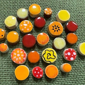 May include: A collection of small, round, flat-topped buttons in various shades of yellow, orange, and red. Some buttons feature decorative patterns including white polka dots, black swirls, and radial lines, while others have solid glossy finishes.