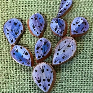 May include: Handmade ceramic cactus pad shapes. Each piece has a blue and purple glaze with black dot and line details, and a brown edge. The pieces are arranged on a green textured surface.