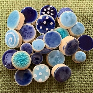 May include: A collection of small, round ceramic cabochons in various shades of blue, including navy, sky blue, and turquoise. The pieces feature diverse finishes such as solid glazes, white polka dots, swirling patterns, and crackled textures.