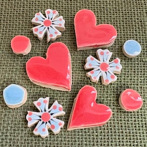 May include: A selection of handmade ceramic tiles in various shapes. The tiles include red hearts, flowers with blue and red accents, and round tiles in red and blue. The tiles are arranged on a textured green surface.