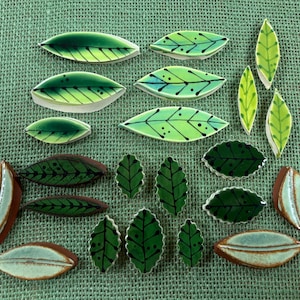 May include: A collection of twenty-two ceramic leaf-shaped tiles in various shades of green, light blue, and brown. The tiles feature hand-painted black vein details, including dots and lines, with some pieces having a smooth, glossy finish.