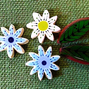 May include: Hand-painted ceramic flower and leaf decorations. Three flower-shaped pieces feature light blue and white petals with a dark blue centre. Two leaf-shaped pieces are green with black vein details and a red-brown border. The pieces are on a green woven surface.