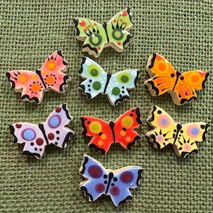 May include: A collection of seven ceramic butterfly-shaped ornaments, each featuring distinct color combinations including lime green, coral pink, light blue, orange, lavender, red, and yellow, all decorated with circular patterns and black outlines.