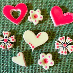 May include: A selection of ceramic decorations, featuring hearts and flowers in pink, white, and black. The hearts are solid and open, and the flowers have dot and petal patterns. The objects are displayed on a green, textured background.