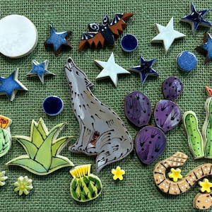 May include: A collection of ceramic tiles featuring a howling wolf, a bat, various cacti, stars, a moon, and a snake. The pieces are glazed in colors including blue, white, green, purple, and brown, with hand-painted details on each individual shape.