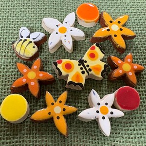 Peut inclure: Divers éléments décoratifs en céramique, dont des fleurs, un papillon, une abeille et des formes rondes. Les pièces présentent des couleurs vives comme l'orange, le jaune, le blanc et le rouge, avec des bordures marron. Sur une surface verte texturée.