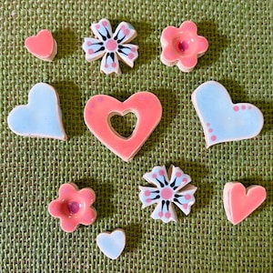 May include: A collection of ceramic heart and flower-shaped decorative pieces in shades of pink, light blue, and white. The pieces are arranged on a textured green surface, showcasing a handmade aesthetic.