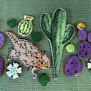 May include: A collection of ceramic mosaic tiles featuring a tan horned lizard, a tall green saguaro cactus, several purple prickly pear cactus pads with black spines, small white flowers with green centers, and small round green accent pieces.