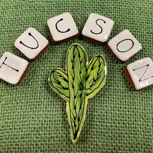 May include: White ceramic tiles spelling out the word "TUCSON" with black lettering, arranged in a semi-circle around a green cactus illustration.