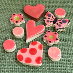 May include: A selection of handmade ceramic decorations in heart, flower, and butterfly shapes. The pieces are predominantly pink with red and black detailing. The items are arranged on a green textured surface. Measurements are in centimetres.