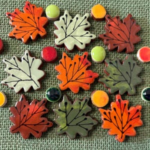 Op de afbeelding: Een verzameling keramische blad- en ronde kralenversieringen in verschillende kleuren. De bladeren zijn in tinten oranje, rood en groen, met zwarte aderdetails. De kralen zijn geel, groen, rood en donkerblauw.