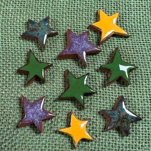 May include: A collection of nine small, five-pointed star-shaped ceramic tiles with a glossy finish. The stars feature various solid and mottled colors, including bright yellow, deep green, purple, and blue, each with a smooth, reflective surface.