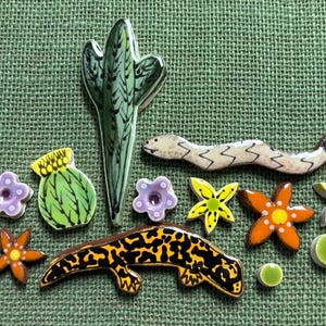 May include: A selection of ceramic tiles with a desert theme, featuring a tall green cactus, a snake, a lizard, flowers, and round green tiles. The tiles are brightly coloured and set against a green textured background.
