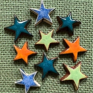 May include: A collection of nine ceramic star-shaped ornaments featuring a glossy, glazed finish. The stars are arranged in various colors, including shades of blue, teal, orange, and light green, each with a smooth, reflective surface texture.