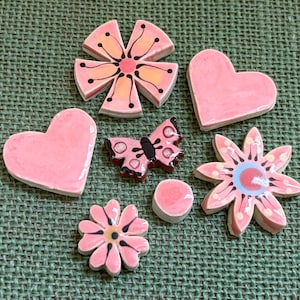 Puede incluir: Una colección de piezas decorativas de cerámica rosa, que incluyen dos corazones, dos formas de flores, una mariposa y un pequeño cilindro. Las piezas tienen detalles negros y están dispuestas sobre una superficie verde texturizada.