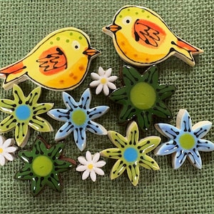 May include: A collection of decorative ceramic shapes including two yellow birds with orange wings, several six-petaled flowers in shades of lime green, light blue, and dark green, and three small white flowers with yellow centers.