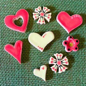 May include: A selection of handmade ceramic tiles in heart and flower shapes. The hearts are mainly pink, some with cutouts or dotted patterns. The flower tiles have pink, black, and green details, all on a green textured background.