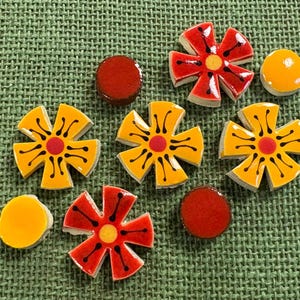 May include: A collection of colourful ceramic tiles in various shapes. The tiles include flower shapes in yellow and red, and round tiles in orange and red. The tiles are arranged on a green textured surface.
