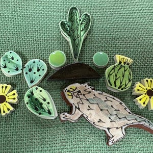 May include: A collection of ceramic mosaic tiles featuring a central saguaro cactus, a horned lizard, two yellow flowers, four green cactus pads, two small green circles, and a dark brown semi-circle piece, all arranged on a textured green surface.