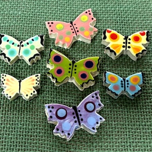 May include: Seven colorful butterfly-shaped ceramic decorations. Each butterfly has a unique design with various colors, including pink, yellow, blue, green, and purple. The butterflies are arranged on a textured green surface.