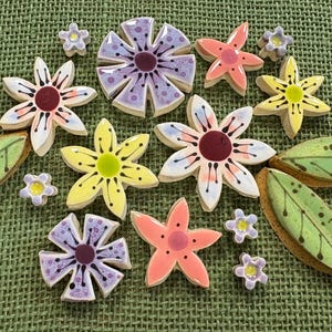 Puede incluir: Una colección de azulejos de cerámica con formas de flores y hojas en varios colores, incluyendo morado, amarillo, rosa y verde. El conjunto presenta flores de varios pétalos con centros oscuros, pequeñas flores de cinco pétalos y hojas alargadas con venas oscuras.