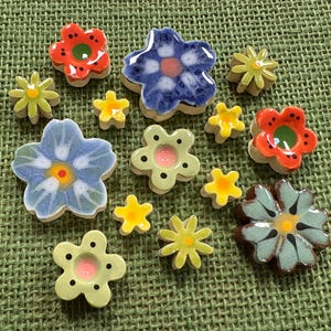 Puede incluir: Una colección de pequeñas formas de flores de cerámica multicolor en varios tamaños. El conjunto incluye flores azules, rojas, verdes y amarillas con acabados brillantes, algunas con puntos pintados o centros, dispuestas en un patrón disperso.