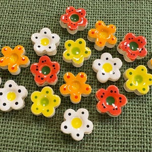 May include: A collection of small, five-petaled flower-shaped buttons in various colors including white with black dots, bright yellow, orange, and red with dark spots. Each button features a smooth, glossy finish and a central circular indentation.