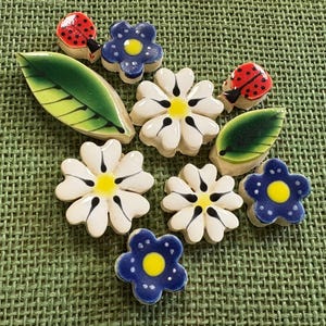May include: A collection of ceramic decorative pieces including two red and black ladybugs, three blue five-petaled flowers with yellow centers, three white six-petaled flowers with yellow and black centers, and two green leaves with dark veins.