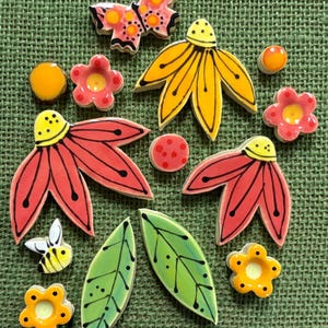 Puede incluir: Una colección de piezas decorativas de cerámica que incluye una mariposa rosa, dos flores rojas grandes, una flor amarilla grande, tres flores pequeñas de cinco pétalos en rosa y amarillo, dos hojas verdes, una abeja pequeña y tres puntos.